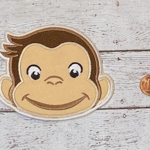May include: Embroidered patch featuring a smiling monkey face with brown hair and accents. The monkey has a light tan face, white eyes, and a brown mouth. The patch is outlined in a white border.