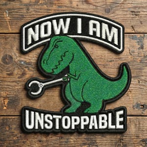 May include: Embroidered patch featuring a green Tyrannosaurus Rex holding a wrench. The patch has the text "NOW I AM" above the dinosaur and "UNSTOPPABLE" below, all in white lettering on a black background.