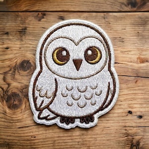 May include: Embroidered owl patch featuring a white body, brown outline, and large yellow eyes. The patch has a detailed design with visible stitching and is set against a wooden background. The owl's design includes a brown beak and feet.
