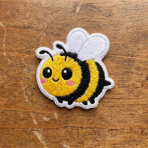 May include: Embroidered bee patch with a smiling face, black and yellow striped body, and white wings. The patch has a white border and is set against a wooden background.