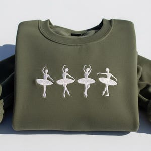May include: Olive green sweatshirt with an embroidered design of four white ballerinas in various dance poses. The ballerinas are wearing tutus. The sweatshirt has a crew neck and long sleeves.