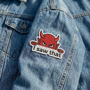 May include: A denim jacket with a patch featuring a red devil with horns and the text "I saw that" in white. The patch is embroidered and sewn onto the blue denim fabric.