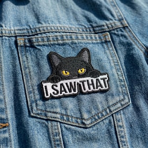 May include: A black cat patch with yellow eyes and the text "I SAW THAT" embroidered on a white banner. The patch is affixed to the pocket of a blue denim jacket.