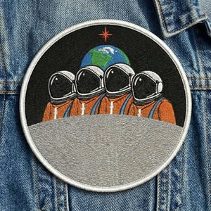 May include: Embroidered patch featuring four astronauts in orange suits and helmets, standing on a gray surface. The background includes a black sky, a blue and green Earth, and a red star. The patch is circular with a white border.