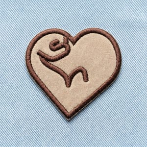May include: A heart-shaped patch with a light tan center and a dark brown embroidered outline. The patch features a stylized brown design within the heart shape, set against a light blue denim background. The patch is likely for clothing or accessories.