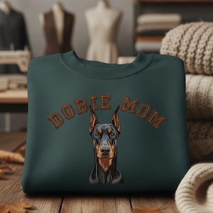 Può includere: Una felpa verde scuro con la scritta ricamata "DOBIE MOM" sopra un ritratto dettagliato di un Dobermann. Il pelo del cane è nero e marrone, con orecchie dritte.