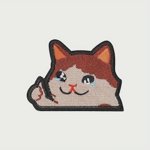 May include: Embroidered patch featuring a cartoon cat with brown and white fur, orange ears, and a red nose. The cat has two blue teardrops and a sad expression. The patch has a black border.