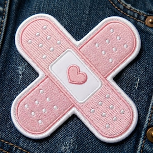 May include: Embroidered pink bandage patch with a white border and a white center featuring a pink heart. The patch is in the shape of a cross and has small white dots.