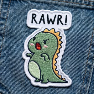 May include: Embroidered patch featuring a green cartoon dinosaur with an angry expression and the word "RAWR!" in a speech bubble. The dinosaur has a yellow spiky back and a white border. The patch is on a blue denim background.