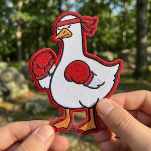 May include: Embroidered patch featuring a white goose wearing a red headband and boxing gloves. The goose has an orange beak and feet, outlined in red. The patch is held by a person's hands against a blurred outdoor background.