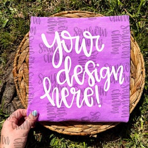 以下が含まれることがあります： 白い筆記体で「your design here!」と書かれた紫色のTシャツ。Tシャツは、背景に草がある編まれたバスケットに表示されています。「Southern Charm」という言葉がシャツに印刷されています。