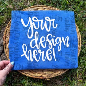 Peut inclure: Un t-shirt bleu roi avec les mots "your design here!" en écriture blanche. Le t-shirt est présenté sur un panier en osier, avec un fond d'herbe verte. Les mots "Southern Charm" sont imprimés en motif sur le t-shirt.