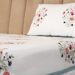 May include: White bed linens with a floral pattern in red, navy, and green. The pillow and sheet feature a repeating design of stylized flowers and foliage. A light blue border trims the pillow and sheet.