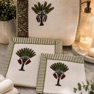 May include: A set of three white waffle-weave towels featuring a green and maroon palm tree motif. The towels have green striped borders, with one towel displaying a floral patterned edge. The fabric has a textured, grid-like surface pattern.