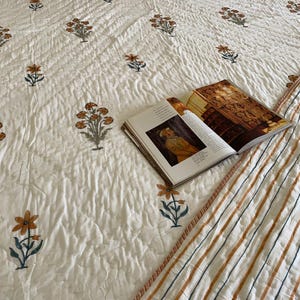 May include: A white quilt with a floral pattern in shades of orange, green, and gray. The quilt has a striped border in orange, blue, and white. An open magazine rests on the quilt.