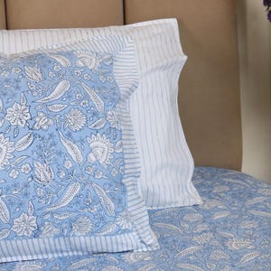 May include: Close-up of a bed with light blue floral patterned bedding and pillows. The pillows have a white and blue striped border. The floral pattern includes white flowers and leaves on a light blue background. The bed is against a neutral-toned headboard.