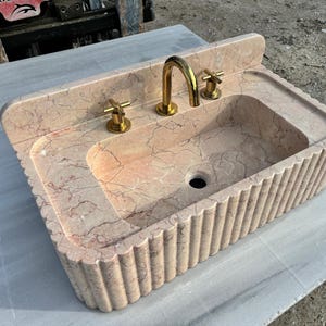 May include: A pink marble sink with gold fixtures. The sink has a rectangular basin with a fluted exterior and a built-in backsplash. The faucet and cross-shaped handles are gold-toned. The sink is made of pink marble with dark veining.