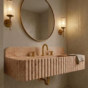 May include: A bathroom vanity with a pink marble sink and gold fixtures. The sink has a fluted design and is paired with a round gold mirror and two wall sconces with cylindrical glass shades. A gray towel rests on the sink.