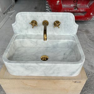 Puede incluir: Un lavabo de m&aacute;rmol blanco con grifos de lat&oacute;n. El lavabo tiene una cubeta rectangular y un salpicadero decorativo. El grifo y los tiradores son de lat&oacute;n. El lavabo est&aacute; sobre una base de madera.