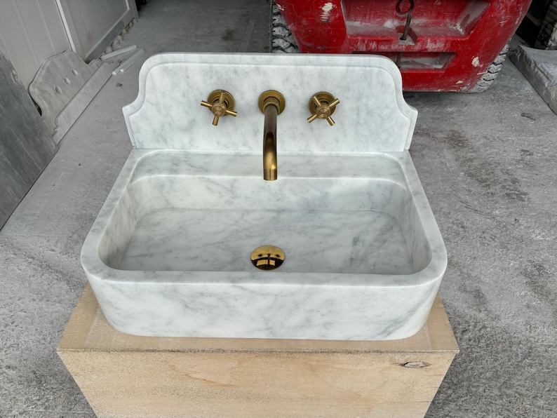 Puede incluir: Un lavabo de m&aacute;rmol blanco con grifos de lat&oacute;n. El lavabo tiene una cubeta rectangular y un salpicadero decorativo. El grifo y los tiradores son de lat&oacute;n. El lavabo est&aacute; sobre una base de madera.