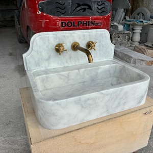 Puede incluir: Un lavabo de m&aacute;rmol blanco con grifos de lat&oacute;n. El lavabo tiene una cubeta rectangular y un protector contra salpicaduras decorativo. El grifo y las manijas son de color dorado. El lavabo se exhibe sobre una caja de madera.