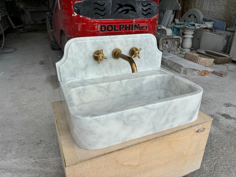 Puede incluir: Un lavabo de m&aacute;rmol blanco con grifos de lat&oacute;n. El lavabo tiene una cubeta rectangular y un protector contra salpicaduras decorativo. El grifo y las manijas son de color dorado. El lavabo se exhibe sobre una caja de madera.