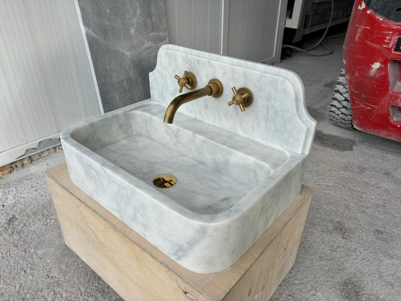 Puede incluir: Un lavabo rectangular de m&aacute;rmol con respaldo curvo y accesorios de lat&oacute;n. El lavabo tiene un patr&oacute;n veteado gris claro y blanco. El grifo y los dos tiradores son de lat&oacute;n. El lavabo est&aacute; sobre una caja de madera.