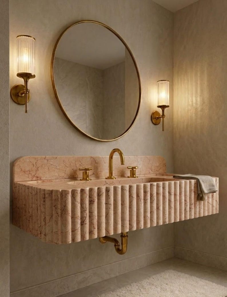 May include: A bathroom vanity with a pink marble sink and gold fixtures. The sink has a fluted design and is paired with a round gold mirror and two wall sconces with cylindrical glass shades. A gray towel rests on the sink.