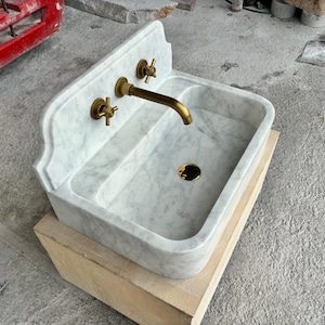 Puede incluir: Un lavabo de m&aacute;rmol blanco con grifos de lat&oacute;n. El lavabo tiene una cubeta rectangular y una parte trasera curvada. El grifo y dos manijas son de lat&oacute;n. El lavabo est&aacute; sobre una base de madera.
