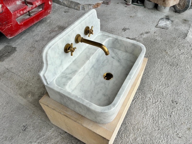 Puede incluir: Un lavabo de m&aacute;rmol blanco con grifos de lat&oacute;n. El lavabo tiene una cubeta rectangular y una parte trasera curvada. El grifo y dos manijas son de lat&oacute;n. El lavabo est&aacute; sobre una base de madera.