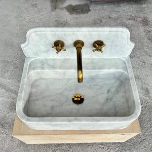 Puede incluir: Un lavabo rectangular de m&aacute;rmol con grifer&iacute;a de lat&oacute;n. El lavabo tiene un patr&oacute;n veteado gris claro y blanco. El grifo y los dos mandos son de lat&oacute;n. El desag&uuml;e es negro y dorado. El lavabo est&aacute; sobre una base marr&oacute;n claro.