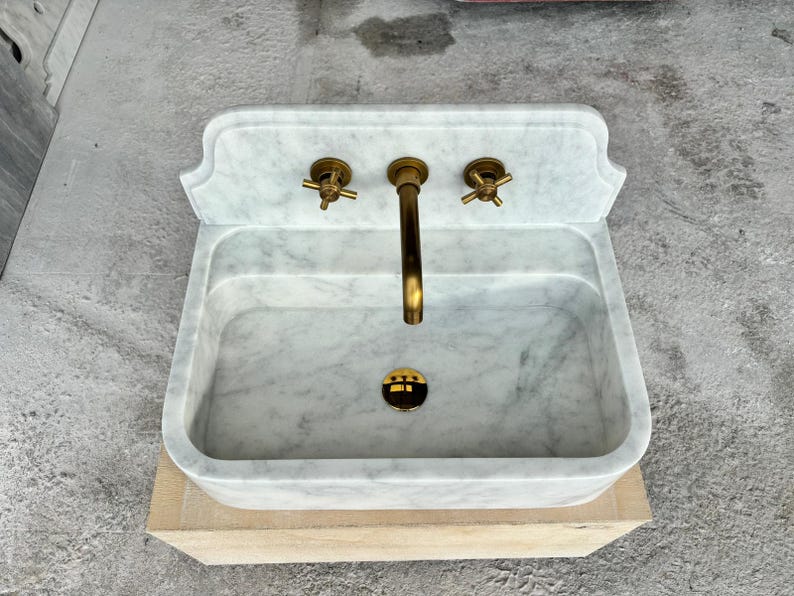 Puede incluir: Un lavabo rectangular de m&aacute;rmol con grifer&iacute;a de lat&oacute;n. El lavabo tiene un patr&oacute;n veteado gris claro y blanco. El grifo y los dos mandos son de lat&oacute;n. El desag&uuml;e es negro y dorado. El lavabo est&aacute; sobre una base marr&oacute;n claro.