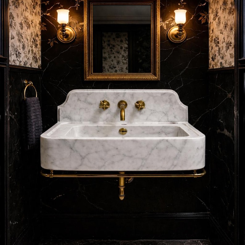 Puede incluir: Un lavabo de ba&ntilde;o de m&aacute;rmol con grifos dorados, colocado contra una pared de m&aacute;rmol negro. El lavabo tiene una cubeta rectangular y un respaldo curvo. Un espejo con marco dorado y apliques de pared est&aacute;n encima del lavabo.