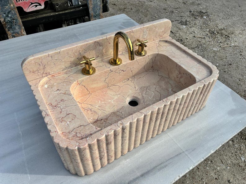 May include: A pink marble sink with gold fixtures. The sink has a rectangular basin with a fluted exterior and a built-in backsplash. The faucet and cross-shaped handles are gold-toned. The sink is made of pink marble with dark veining.