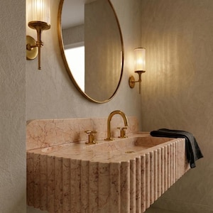 May include: A bathroom scene featuring a pink marble sink with gold fixtures, an oval gold-framed mirror, and two wall sconces with cylindrical glass shades. A dark gray towel hangs on the sink. The overall design is modern and luxurious.