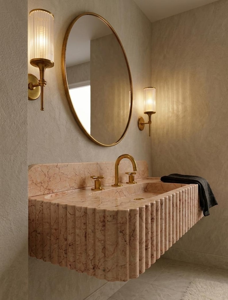 May include: A bathroom scene featuring a pink marble sink with gold fixtures, an oval gold-framed mirror, and two wall sconces with cylindrical glass shades. A dark gray towel hangs on the sink. The overall design is modern and luxurious.