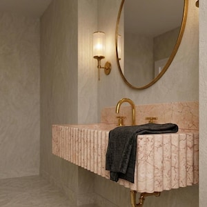 May include: A modern bathroom sink with a pink marble countertop and a gold faucet. A black towel rests on the sink. A gold-framed oval mirror and a gold sconce with a ribbed glass shade are mounted on the wall.