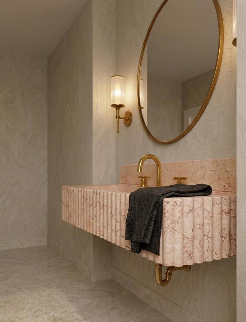 May include: A modern bathroom sink with a pink marble countertop and a gold faucet. A black towel rests on the sink. A gold-framed oval mirror and a gold sconce with a ribbed glass shade are mounted on the wall.