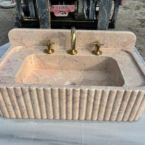May include: A rectangular, peach-colored marble sink with a fluted base and a built-in backsplash. The sink has a central basin and two side ledges. It features a gold faucet and two gold handles. The sink's dimensions are approximately 36 inches wide, 24 inches deep, and 12 inches high.