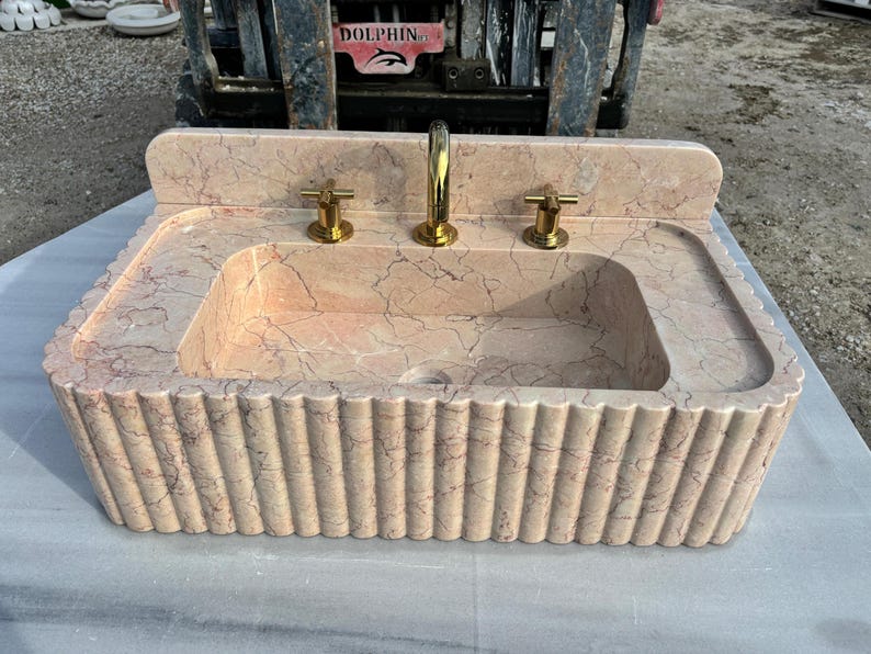 May include: A rectangular, peach-colored marble sink with a fluted base and a built-in backsplash. The sink has a central basin and two side ledges. It features a gold faucet and two gold handles. The sink's dimensions are approximately 36 inches wide, 24 inches deep, and 12 inches high.