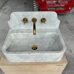 Puede incluir: Un lavabo rectangular de m&aacute;rmol con una placa trasera, con un grifo de lat&oacute;n y dos manijas de lat&oacute;n. El lavabo tiene un patr&oacute;n veteado blanco y gris. El lavabo est&aacute; sobre una caja de madera.