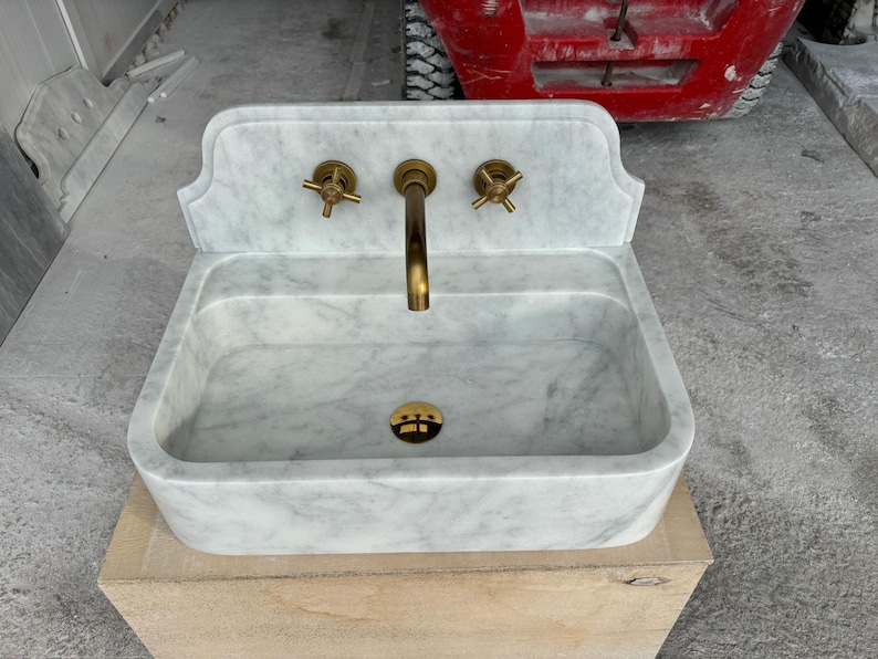 Puede incluir: Un lavabo rectangular de m&aacute;rmol con una placa trasera, con un grifo de lat&oacute;n y dos manijas de lat&oacute;n. El lavabo tiene un patr&oacute;n veteado blanco y gris. El lavabo est&aacute; sobre una caja de madera.