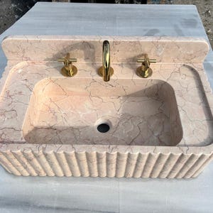 May include: A rectangular, peach-colored marble sink with a scalloped edge. The sink has a built-in backsplash and features gold-colored faucets. The sink basin has a central drain hole.