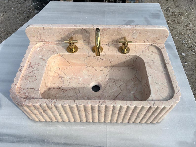 May include: A rectangular, peach-colored marble sink with a scalloped edge. The sink has a built-in backsplash and features gold-colored faucets. The sink basin has a central drain hole.