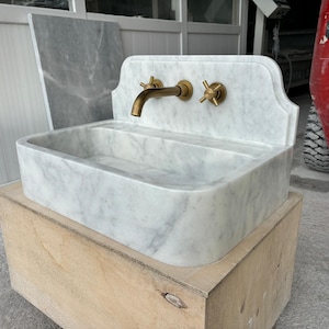 Puede incluir: Un lavabo de m&aacute;rmol blanco con grifos de lat&oacute;n, que incluyen un grifo y dos manijas en forma de cruz. El lavabo tiene una cubeta rectangular y est&aacute; colocado sobre una base de madera. El m&aacute;rmol tiene vetas grises.