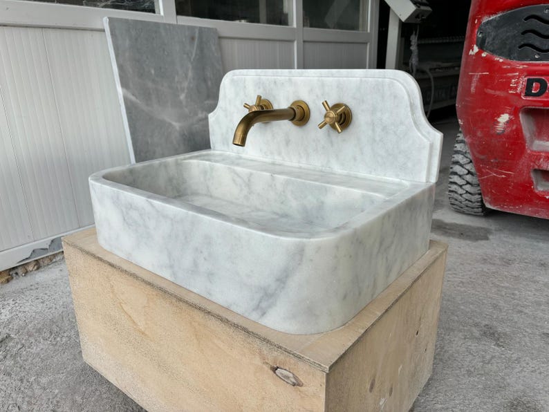 Puede incluir: Un lavabo de m&aacute;rmol blanco con grifos de lat&oacute;n, que incluyen un grifo y dos manijas en forma de cruz. El lavabo tiene una cubeta rectangular y est&aacute; colocado sobre una base de madera. El m&aacute;rmol tiene vetas grises.