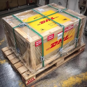 Puede incluir: Una gran caja de madera asegurada con correas verdes y envoltura de pl&aacute;stico transparente, apoyada sobre una paleta de madera. La caja presenta la marca DHL amarilla y roja y etiquetas "FR&Aacute;GIL", indicando env&iacute;o y manipulaci&oacute;n.