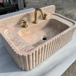May include: A rectangular, peach-colored marble sink with a fluted exterior. The sink features a built-in backsplash, a central basin, and a gold-colored faucet and handles. The marble has natural veining.