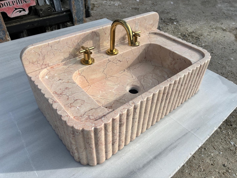 May include: A rectangular, peach-colored marble sink with a fluted exterior. The sink features a built-in backsplash, a central basin, and a gold-colored faucet and handles. The marble has natural veining.