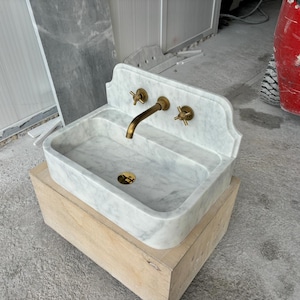 Puede incluir: Un lavabo rectangular de m&aacute;rmol con grifer&iacute;a de lat&oacute;n, que incluye un grifo y dos manijas. El lavabo es de color gris claro con vetas grises m&aacute;s oscuras y se asienta sobre una base de madera de color claro. El grifo y las manijas tienen un acabado dorado.
